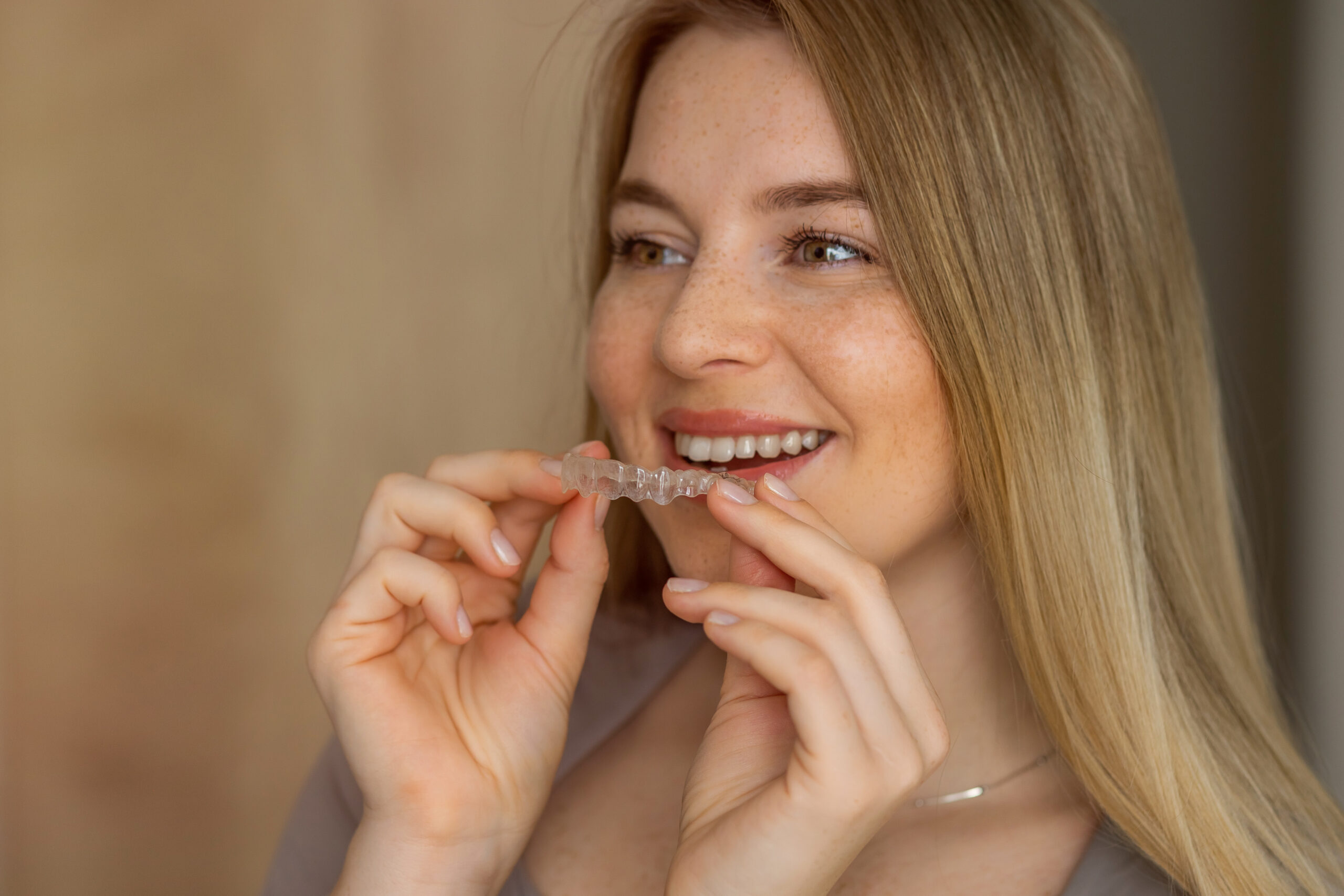 A young woman smiles while holding a clear dental aligner, showcasing modern orthodontic care. Perfect teeth correction and aesthetic restoration aid concept.