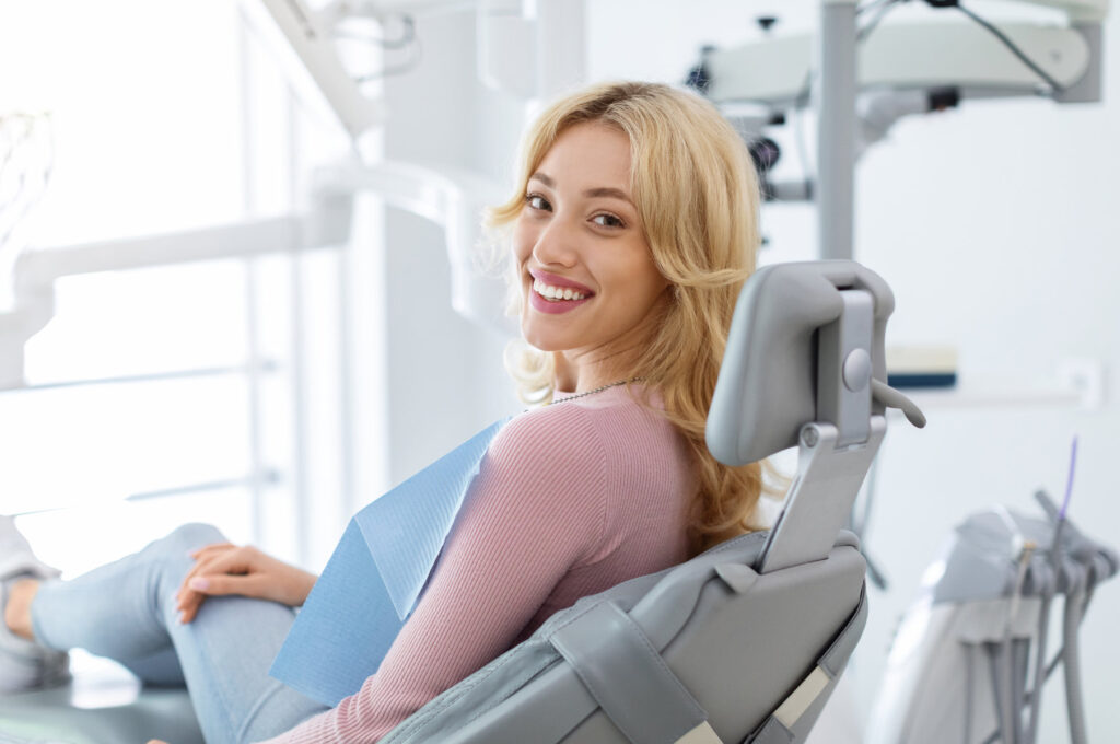 Smiling and relaxed young woman sitting at dental chair in modern clinic, waiting for her dentist. Happy attractive blonde lady attending luxury dental clinic, bacl view, copy space