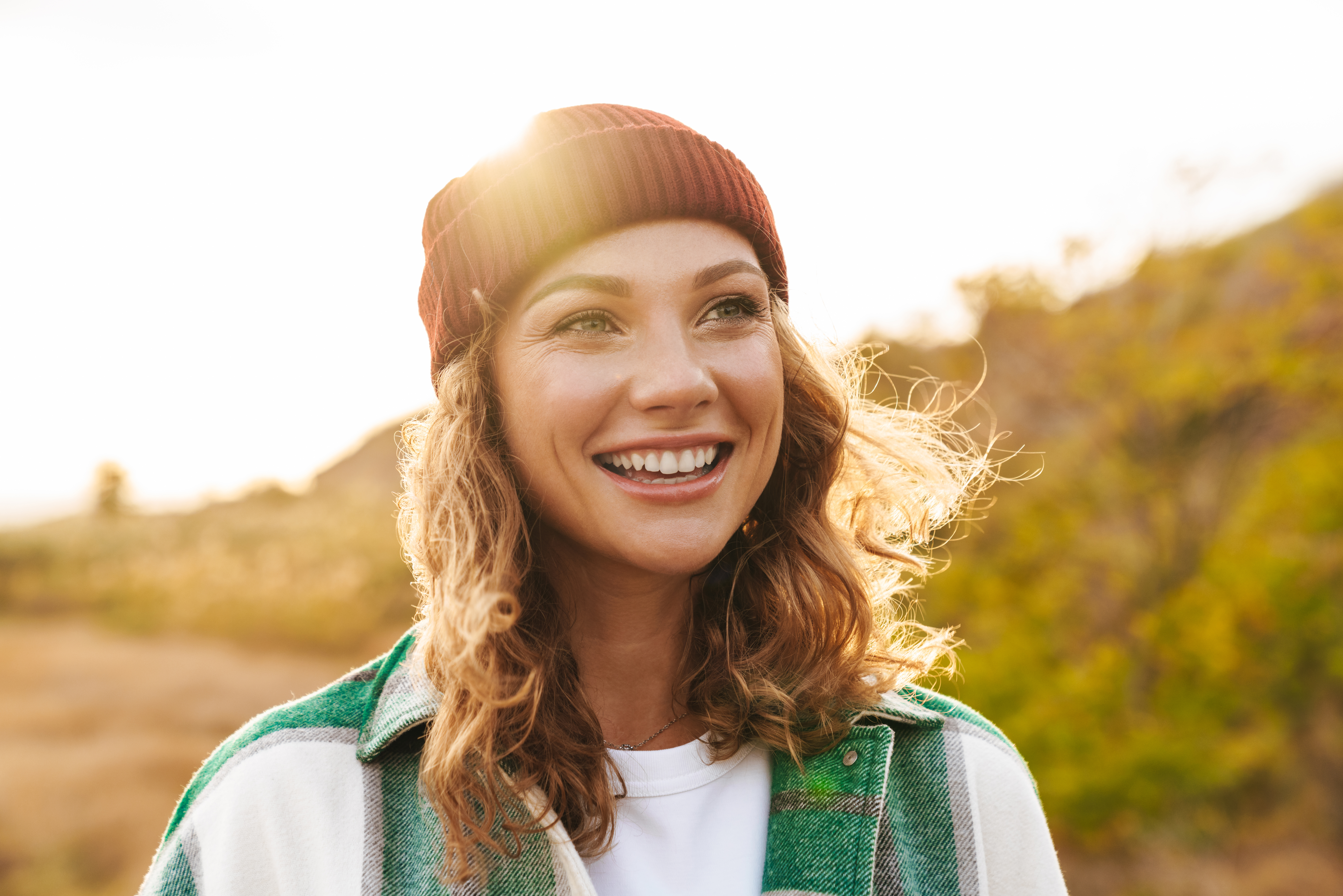 Image of joyful young caucasian woman wearing hat and plaid shirt smiling while walking outdoors