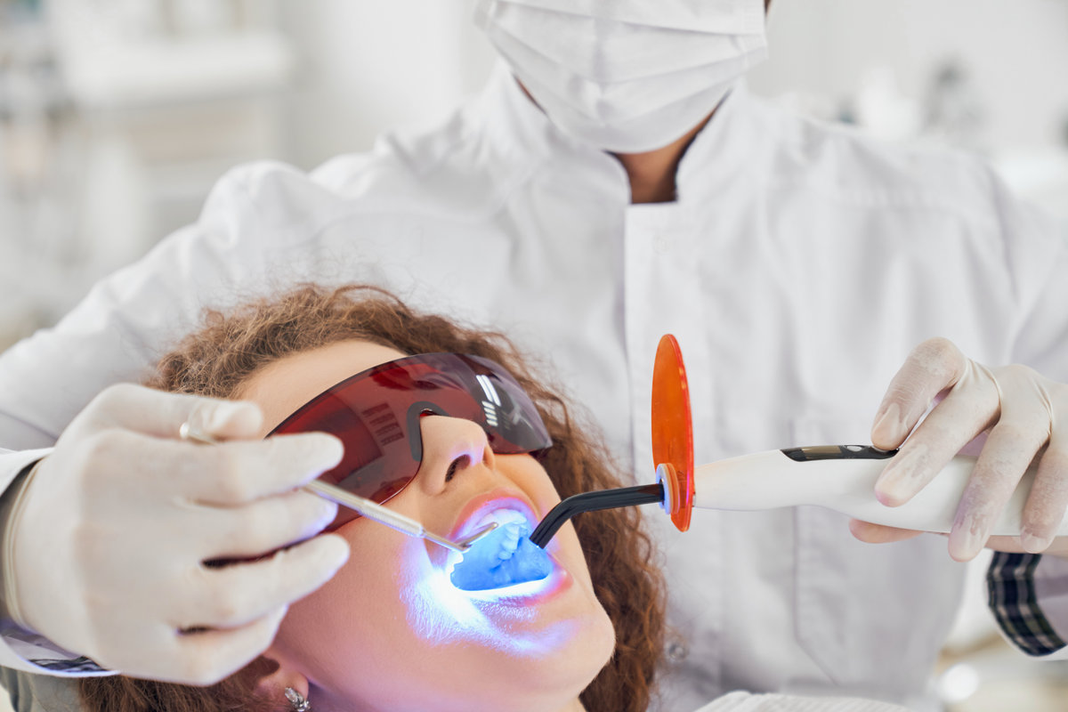 Attractive lady with curly hair lying in dental chair in protective glasses with opened mouth while while male doctor in white glasses doing whitening procedure. Concept of dental care