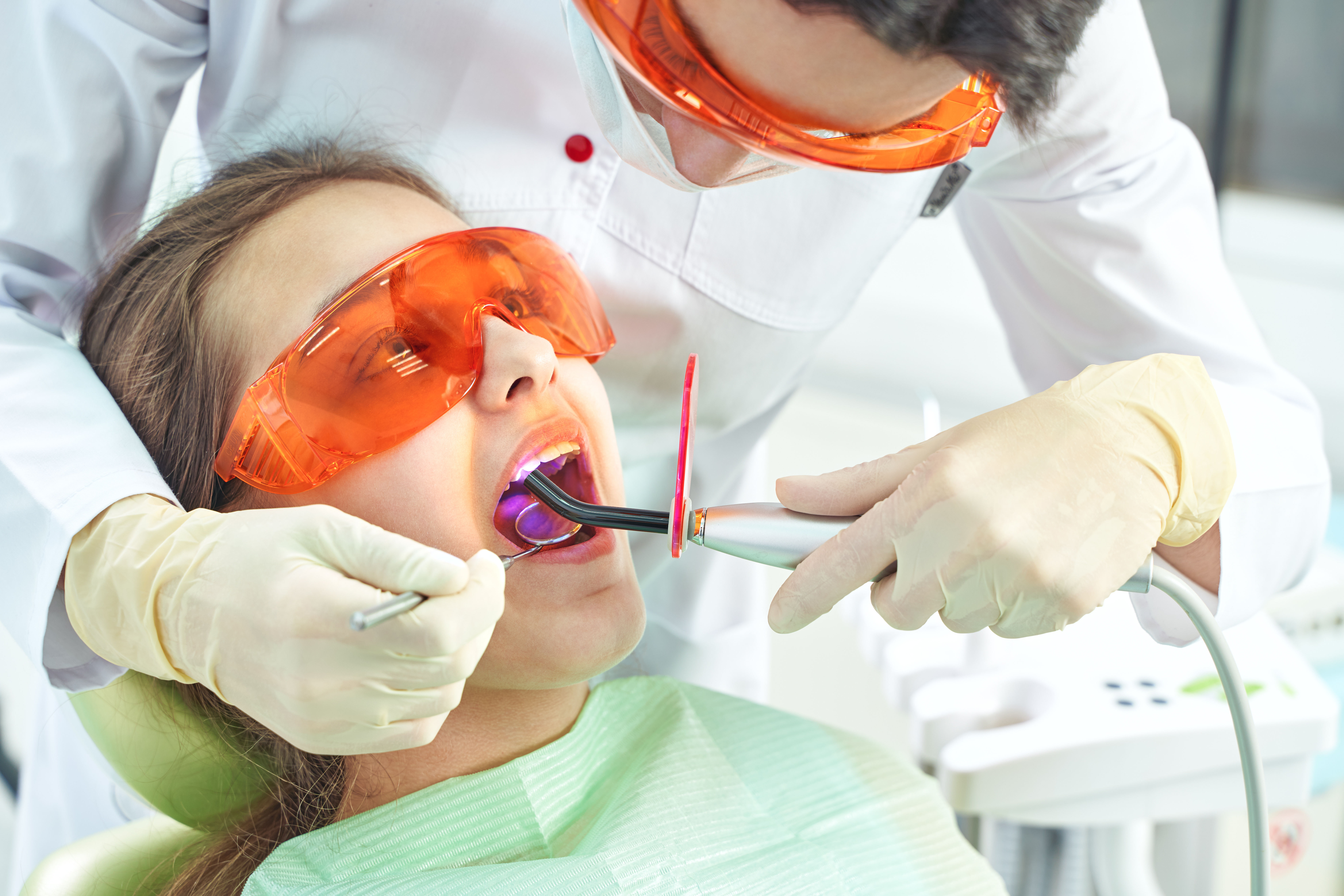 Girl child at the doctor. Dentist places a filling on a tooth with dental polymerization lamp in oral cavity. over clinic background.