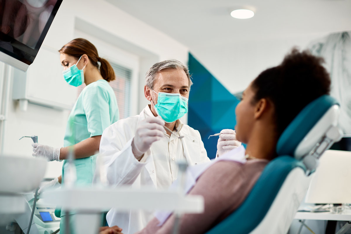 Smiling dentist communicating with African American woman while checking her teeth during dental procedure at dentist's office.
