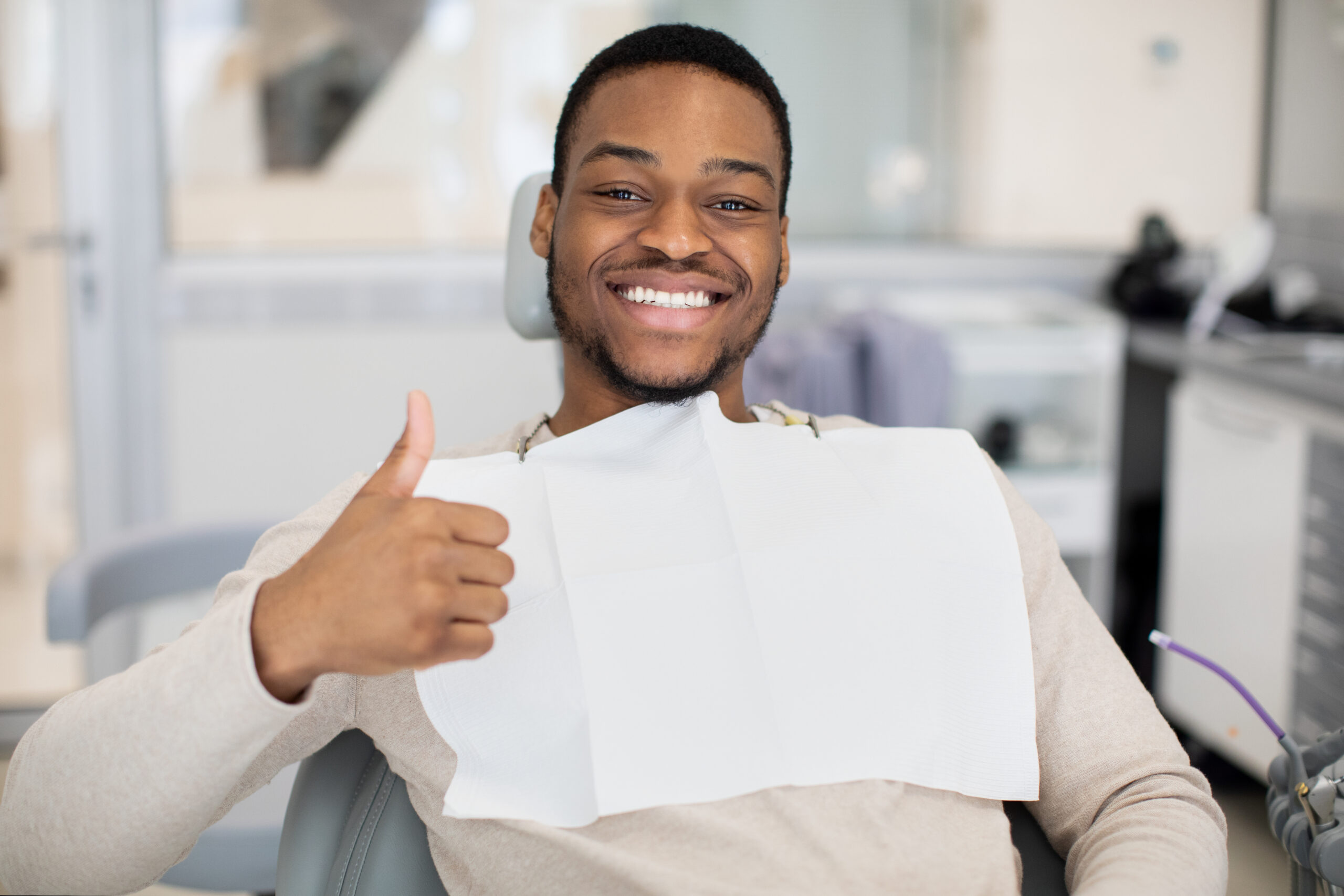 Happy young black man sitting in dentist chair, showing thumb up and smiling at camera. Handsome african american male patient enjoying quality dental services in modern stomatological clinic