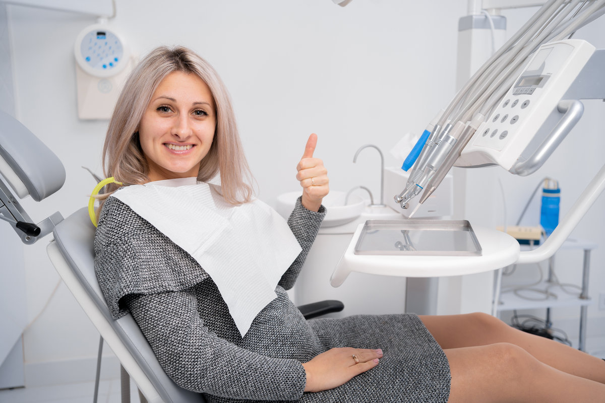 Visit to the dental clinic. Young positive female client showing thumbs up and smiling during dental checkup and consultation in dentist office. copy space.