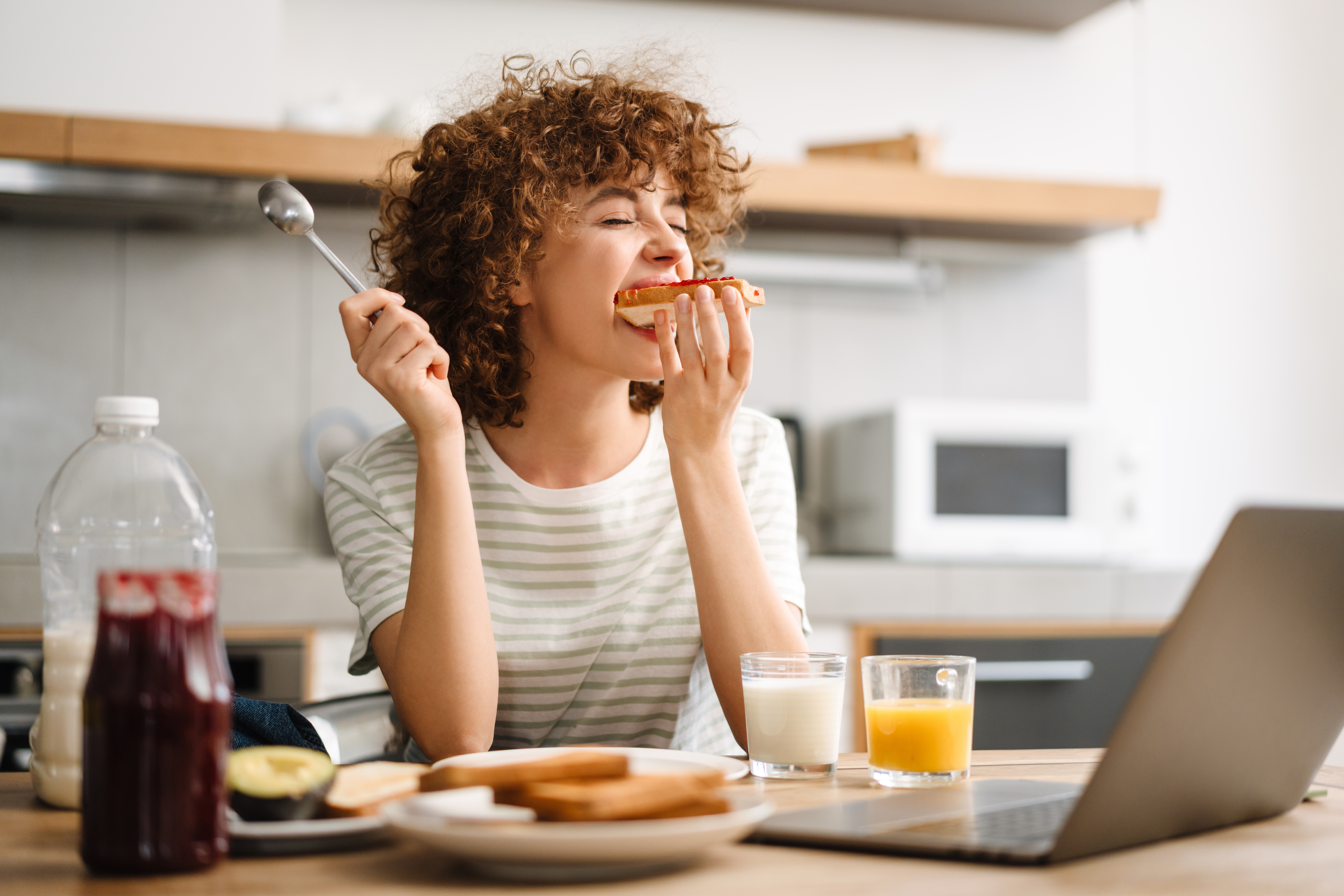 Smiling young woman using laptop while having breakfast at home kitchen