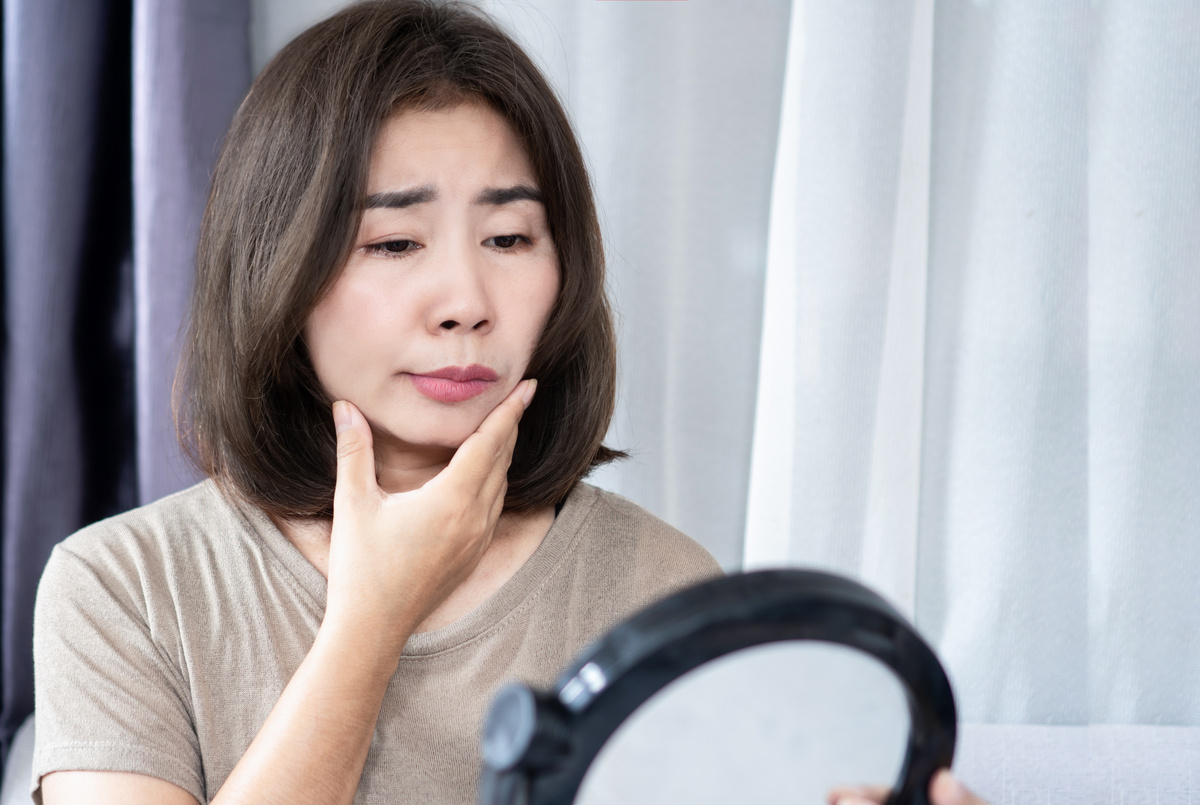 Asian woman holding her face and looking in the mirror , signs of Bell’s palsy or facial paralysis. Her mouth appears drooping, showing facial weakness possibly caused by viral infection, nerve damage